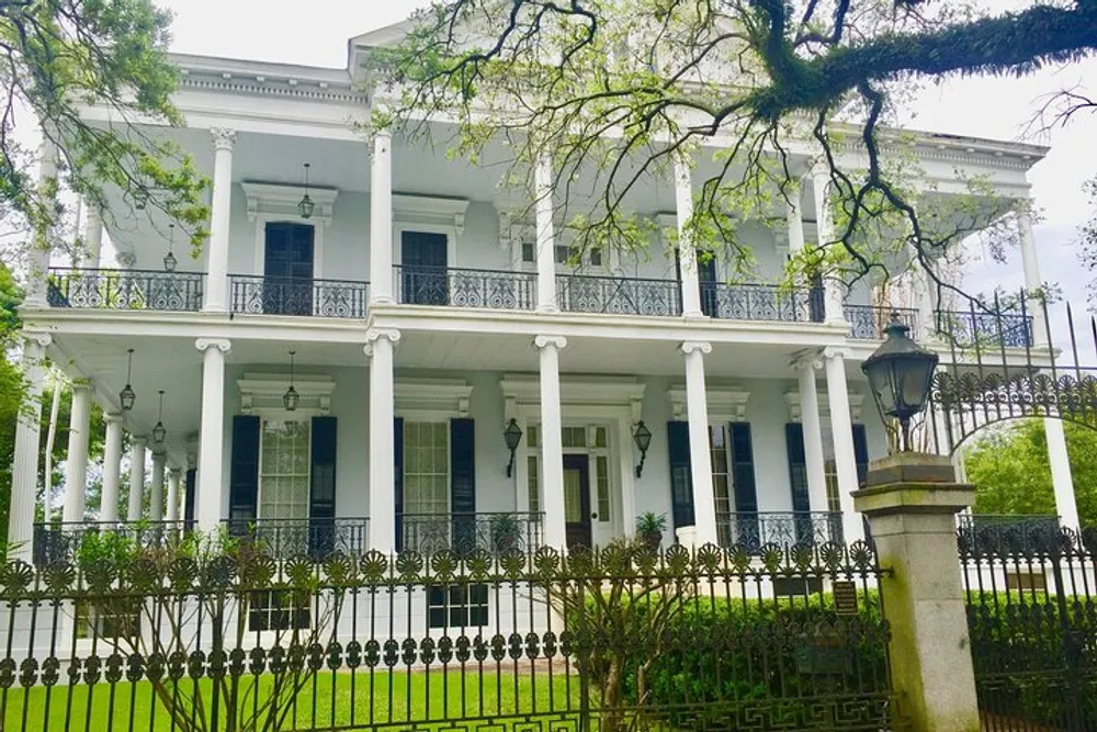 The image shows a grand white two-story mansion with tall columns ornate ironwork on the balconies and surrounded by a lush garden and an ornamental fence