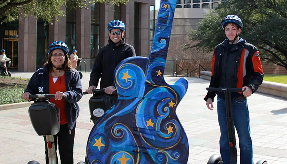 Three people wearing helmets are posing with Segways next to an oversized artistically decorated guitar sculpture