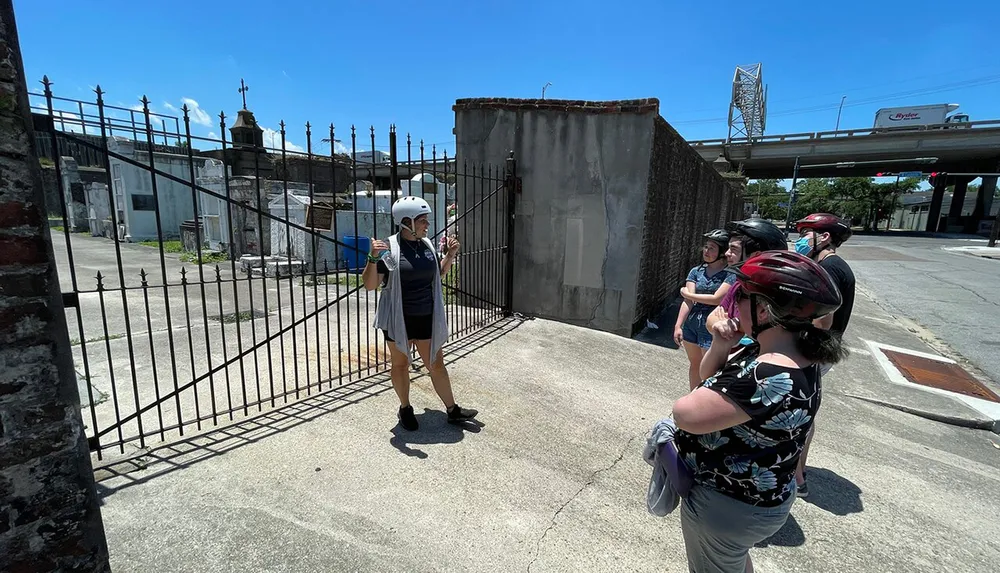 A tour guide wearing a helmet is speaking to a small group of attentive visitors also wearing helmets in an urban setting near a gate with a bridge in the background