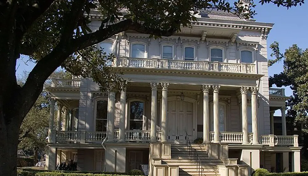 The image shows a large historic-looking two-story mansion with a prominent front balcony and stately columns partially obscured by a tree in the foreground