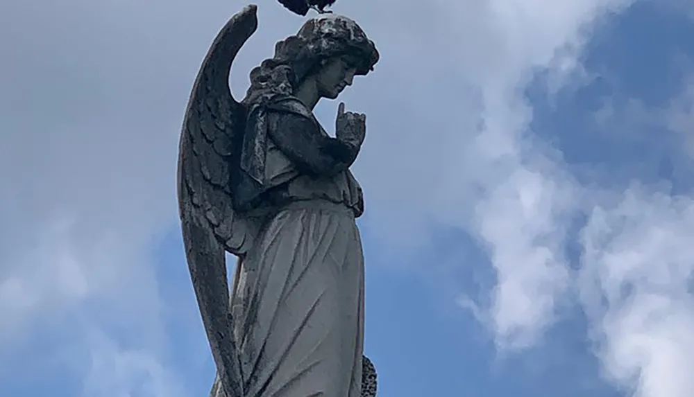 A statue of an angel with folded wings and a contemplative expression stands against a sky with clouds