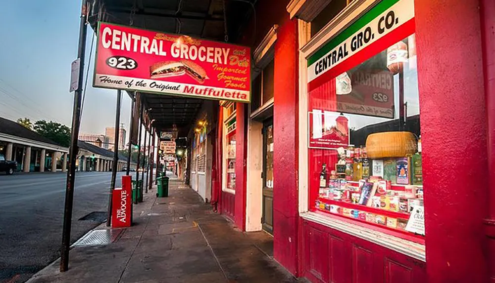 The image shows the vibrant red exterior of Central Grocery a famous deli in New Orleans claiming to be the home of the original muffuletta sandwich set against the backdrop of a quiet street at dusk or dawn