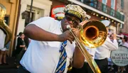 A trombone player performs enthusiastically on the street alongside other brass band members during what appears to be a lively parade or street festival.