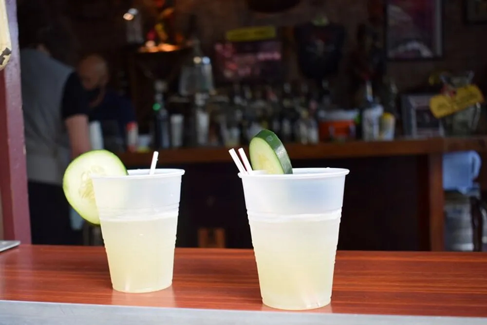 Two plastic cups filled with a light-colored beverage each garnished with a cucumber slice are sitting on a bar counter with a blurred background of a bar setting
