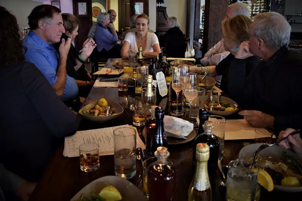 A group of people are engaged in conversation while sitting around a table that is set with food drinks and used dishes indicating a shared meal in a restaurant setting