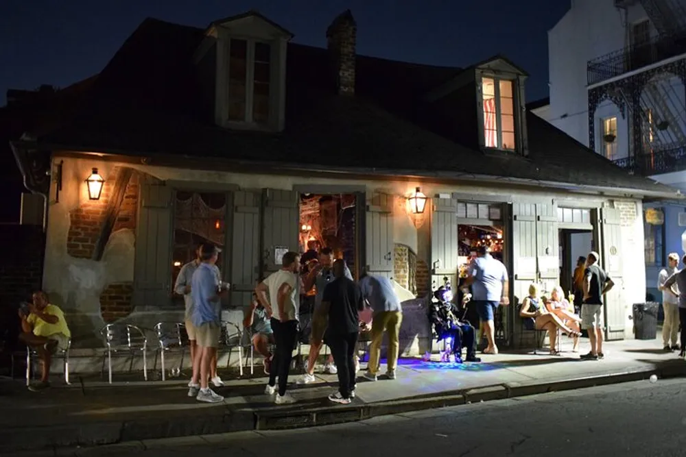 A group of people socialize outside a quaint building with warm lighting at night suggesting a lively and casual evening atmosphere
