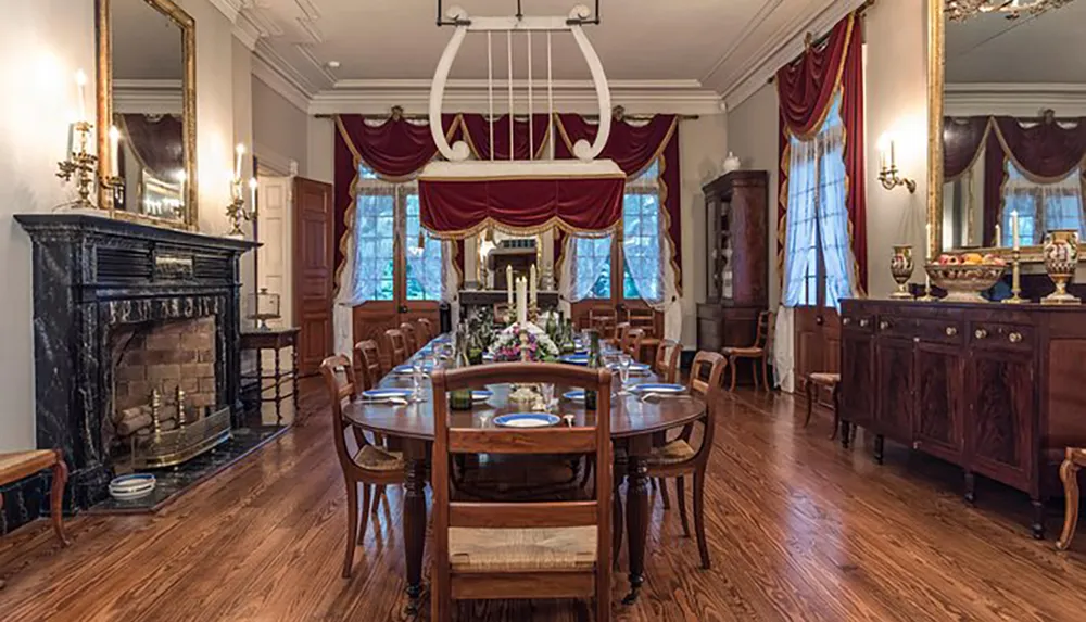 The image shows an elegantly appointed traditional dining room with a long wooden table set for a meal ornate furniture a fireplace and draped windows