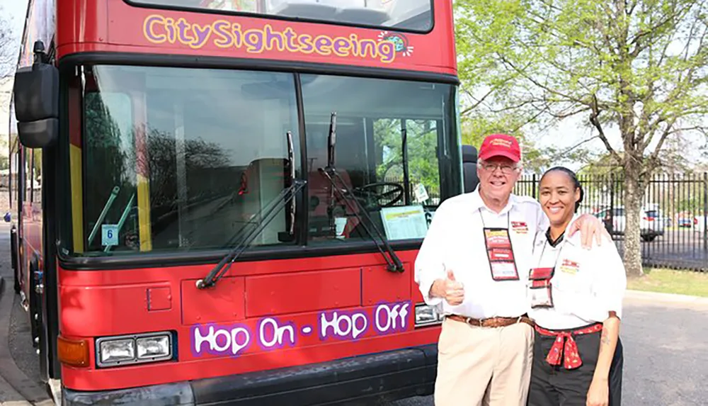 A man and a woman both wearing uniforms and name tags are posing for a photo in front of a red City Sightseeing double-decker bus with a Hop On - Hop Off sign