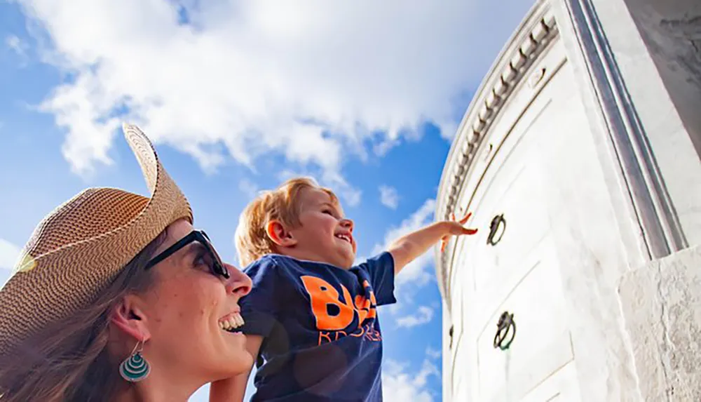A child on the left is excitedly pointing at something out of frame while a woman wearing a straw hat looks on with a smile both set against a backdrop of blue sky and a white tower with Roman numerals