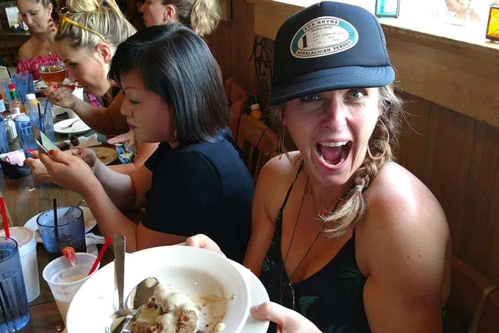 A woman in a trucker hat with an excited expression is holding up a dessert plate at a busy restaurant while another person is eating looking away from the camera