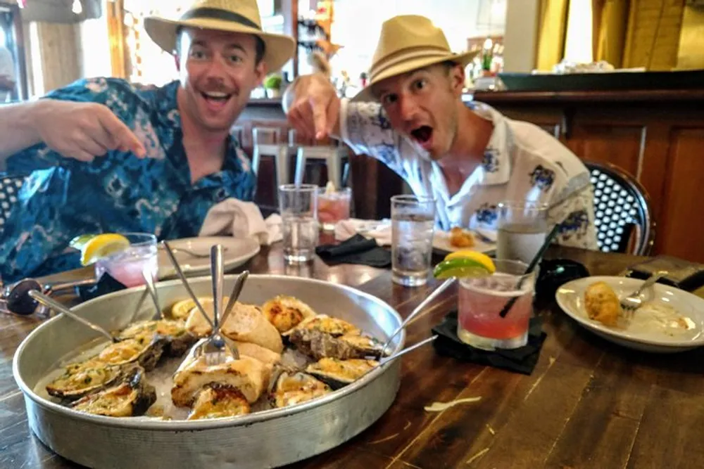 Two people wearing straw hats and casual shirts are enthusiastically pointing at a tray of oysters in a restaurant setting