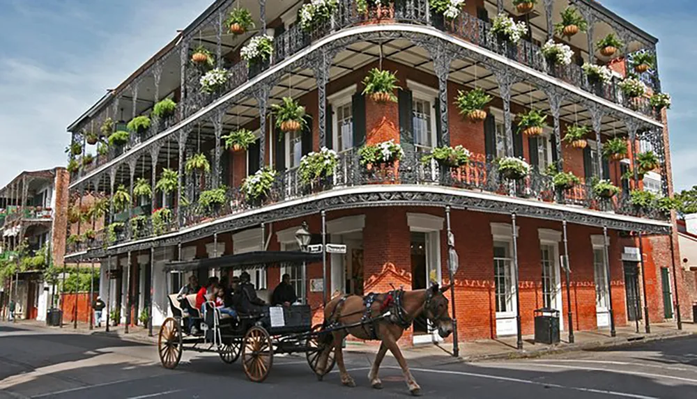 A horse-drawn carriage passes by a traditional brick building with intricate iron balconies adorned with hanging plants evoking the historic charm of the French Quarter in New Orleans