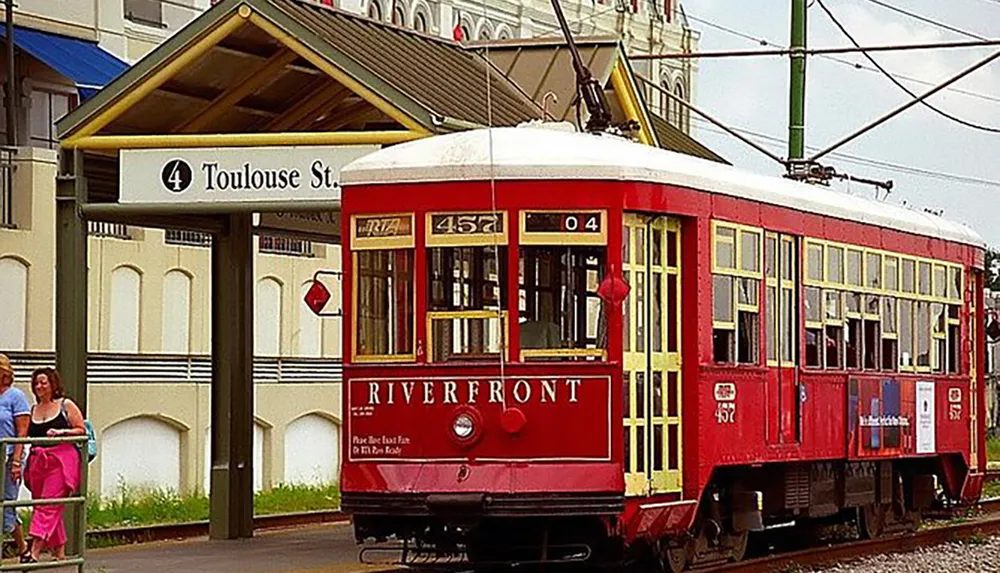 A red and yellow streetcar marked RIVERFRONT is stationed by a stop sign under the Toulouse St sign as passengers wait nearby