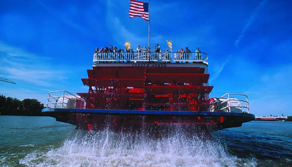 A classic red paddle steamer with passengers on board churning through the water under a clear blue sky proudly flying an American flag at its stern