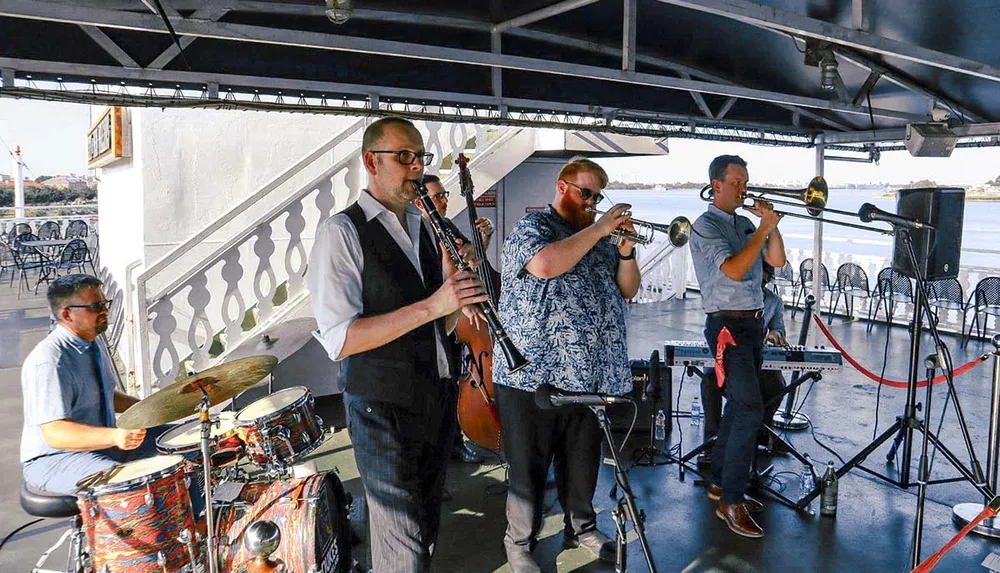 The image shows a live band performing outdoors with members playing drums a double bass clarinets trumpets and a keyboard