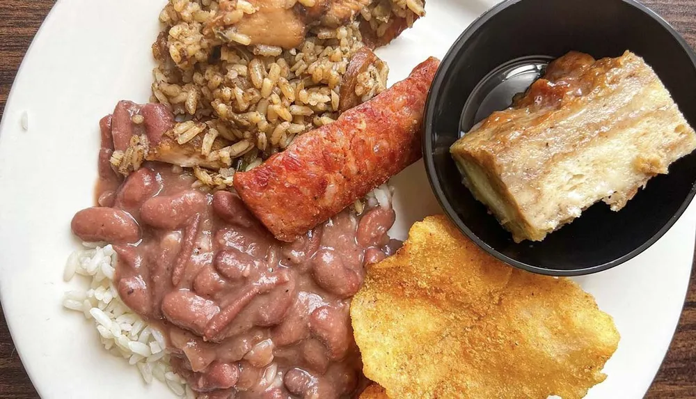 The image shows a plate of food consisting of red beans over rice smoked sausage dirty rice a piece of cornbread and what appears to be a slice of pie suggestive of a Southern or Cajun-style meal