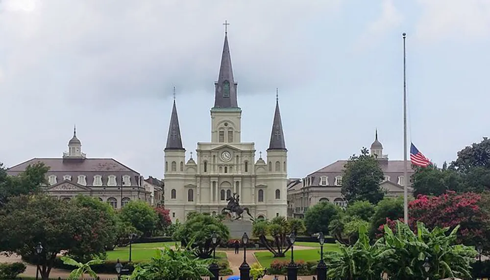 The image shows the iconic St Louis Cathedral with its three spires fronting Jackson Square in New Orleans under an overcast sky with a statue well-kept greenery and an American flag in the foreground
