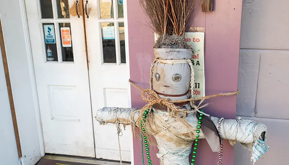 The image shows a weathered and mummified effigy decorated with Mardi Gras beads mounted by a purple wall next to a white door