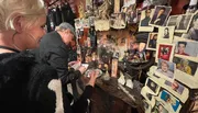 The image shows two people engaging in some kind of activity at a table adorned with various memorabilia and photographs, giving the impression of a shrine or an eclectic collection.