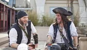 Two people dressed as pirates are laughing and enjoying themselves at a table with a bottle and pirate-themed items.