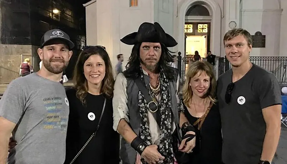 Four smiling individuals pose for a photo with a person dressed dramatically as a pirate resembling a character similar to Captain Jack Sparrow from the Pirates of the Caribbean movies against a backdrop of white classical architecture at night