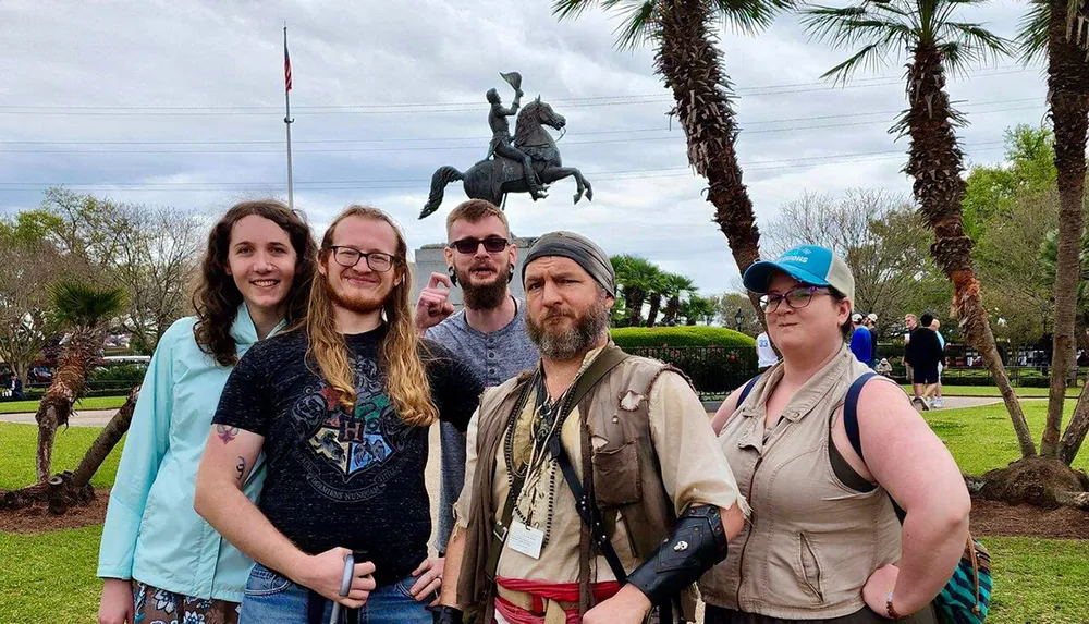 A group of five individuals are posing for a photo in front of a statue of a horse-mounted figure with some of them smiling and one person dressed in pirate-like attire set against a backdrop of palm trees and a cloudy sky