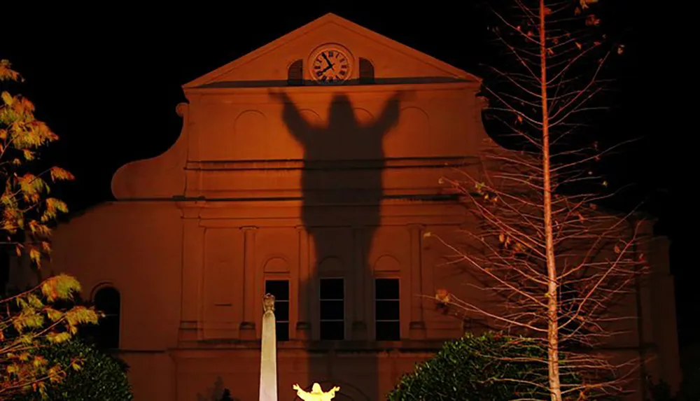 The image shows the shadow of a statue cast on the facade of a building creating an illusion of a large human figure with outstretched arms against the structure at night