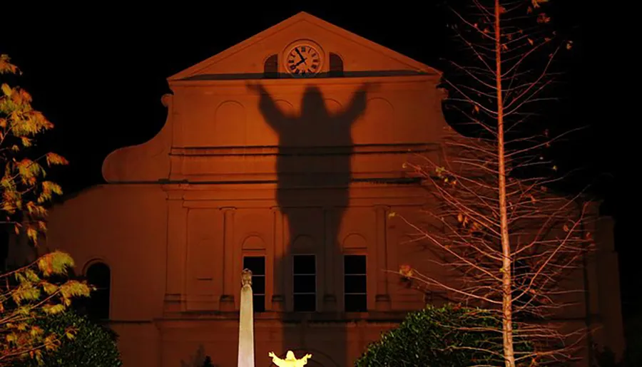 The image shows the shadow of a statue cast on the facade of a building, creating an illusion of a large human figure with outstretched arms against the structure at night.