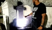 A person wearing a t-shirt with a skull design is pointing at an inscription on a tombstone during nighttime.