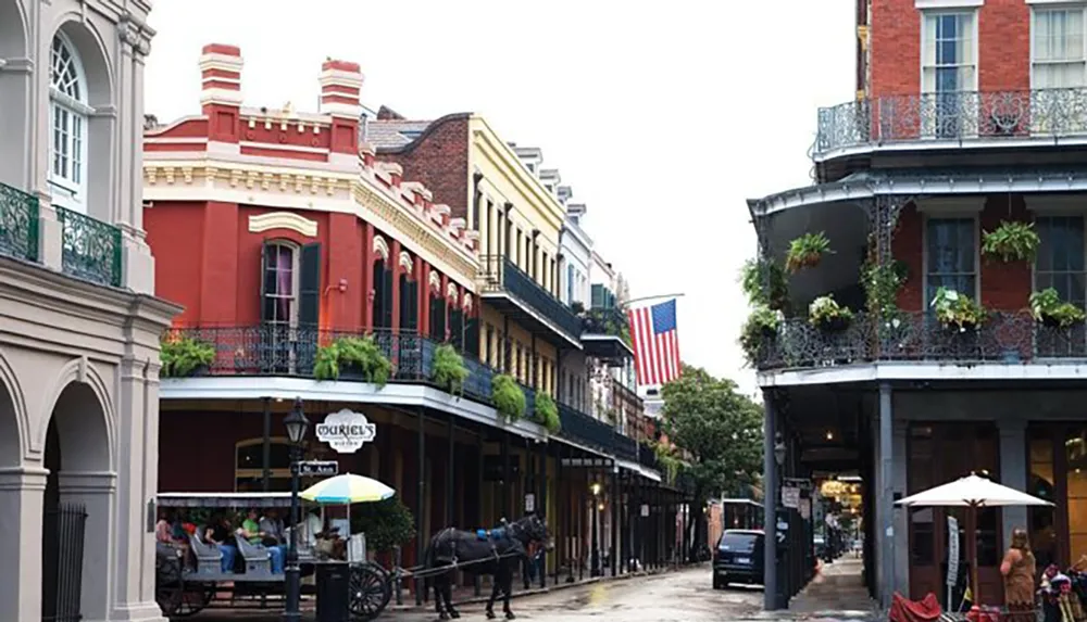 The image shows a street scene with historic multi-storied buildings sporting wrought iron balconies and an American flag patrons dining at sidewalk cafes and a horse-drawn carriage reminiscent of the French Quarter in New Orleans