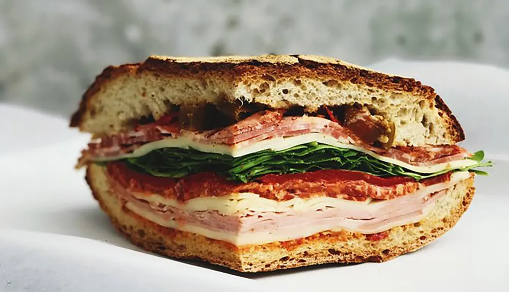 The image shows a thick sandwich with multiple layers of meats cheese tomato and leafy greens between two slices of bread