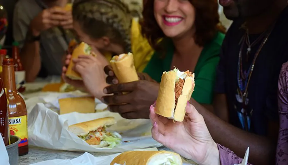 A group of people is enjoying sandwiches together with condiments on the table and one woman smiling at the camera holding a half-eaten sandwich