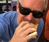 A man wearing sunglasses is biting into a sandwich outdoors