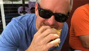 A man wearing sunglasses is biting into a sandwich outdoors.