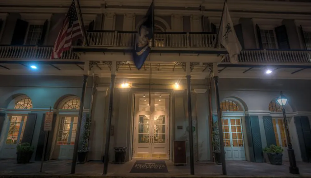 The image depicts the atmospheric facade of a two-story building at night with an illuminated entrance flanked by American and state flags above a balcony characteristic of traditional architecture in New Orleans
