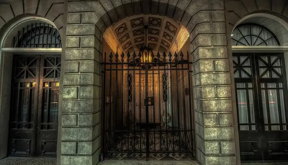 The image shows a dimly lit stone archway entrance with an elegant iron gate and warm light emanating from a hanging lantern inside