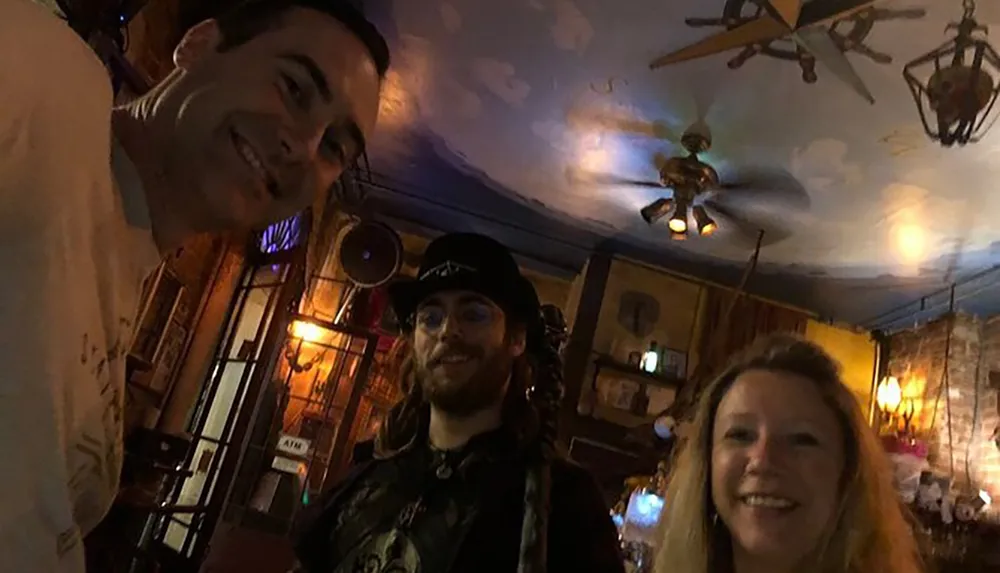 Three people are posing for a selfie with warm smiles in an eclectic dimly-lit room adorned with various hanging objects and a painted ceiling