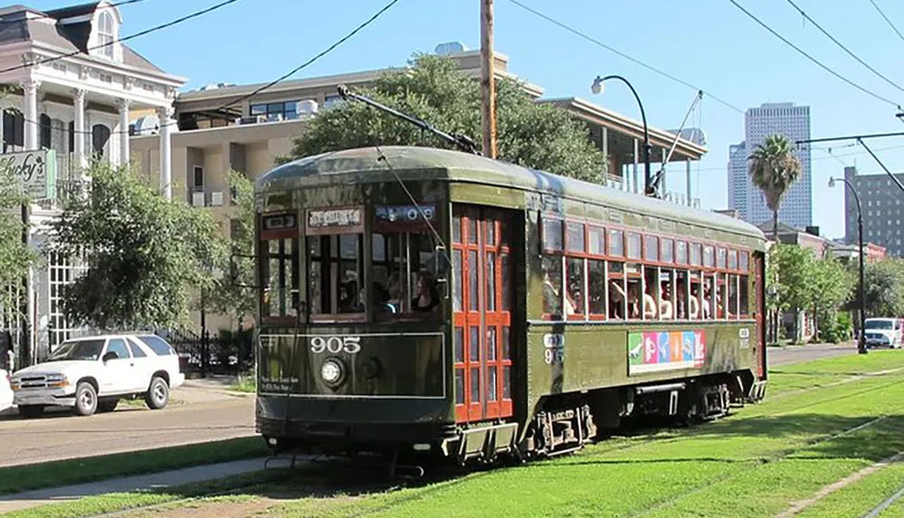 A green vintage streetcar is traveling along a city track with passengers visible inside set against a backdrop of urban buildings and clear skies