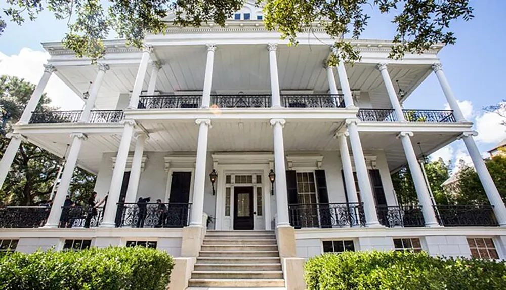 The image shows a grand two-story white house with iconic Southern architecture complete with large columns balconies and a lush front garden