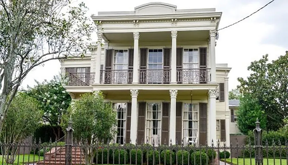 This is an image of a two-story house with a columned front porch and ornate iron balcony railings characteristic of historic southern architecture