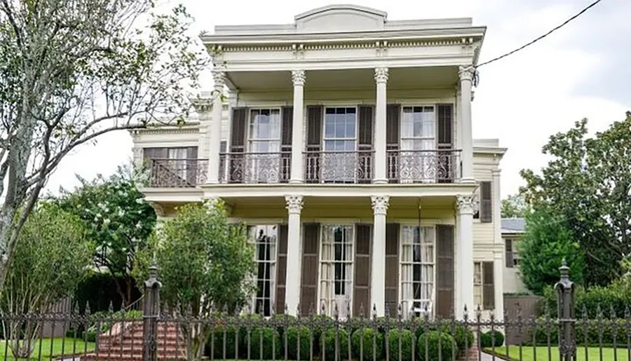 This is an image of a two-story house with a columned front porch and ornate iron balcony railings, characteristic of historic southern architecture.