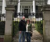 A smiling couple is posing in front of an ornate gate that leads to an elegant white house with black shutters and classic pillars