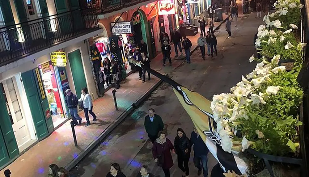 This image shows a bustling pedestrian street at night with people walking past shops and brightly lit establishments adorned with a balcony lined with white flowers