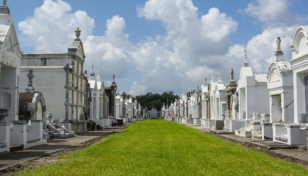 The image features a symmetrical view down a lush green pathway flanked by rows of ornate above-ground white tombs under a partly cloudy sky