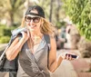 A smiling woman in casual clothing with headphones and sunglasses is walking outdoors while holding a smartphone