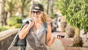 A smiling woman in casual clothing with headphones and sunglasses is walking outdoors while holding a smartphone.