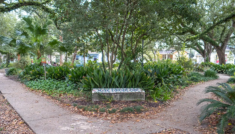 A tranquil park pathway flanked by lush greenery and trees with a concrete sign reading ALICE FORYTH PARK