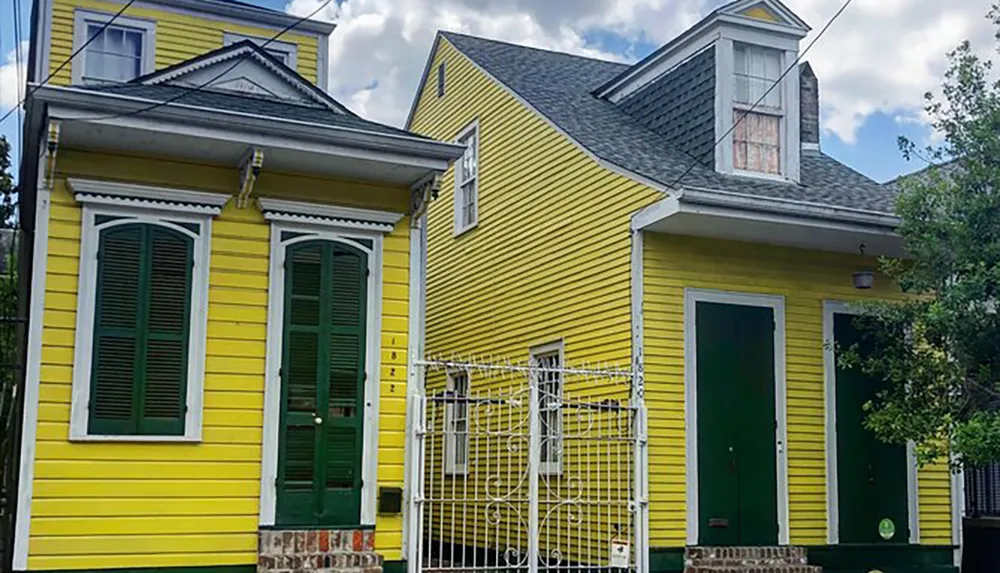 The image shows a vibrant yellow house with green shutters and a front gate possibly located in a warm climate or historic neighborhood