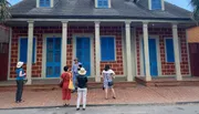 A group of people is listening to a person who appears to be giving a guided tour in front of a distinctive building with blue shutters.