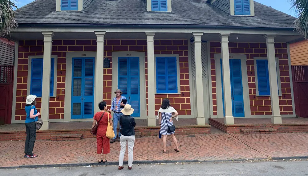 A group of people is listening to a person who appears to be giving a guided tour in front of a distinctive building with blue shutters